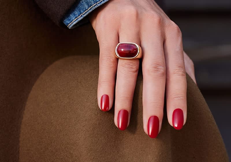 Hände mit roten, glänzenden Fingernägeln liegen auf braunem Stoff, rechter Mittelfinger trägt großen goldenen Ring mit ovalem roten Schmuckstein