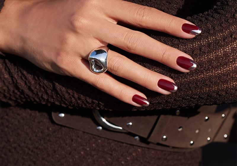 Hand mit dunkelrotem French-Nageldesign und silberner Spitze, trägt großen silbernen Ring und liegt auf braunem Stoff mit sichtbarem braunen Gürtel