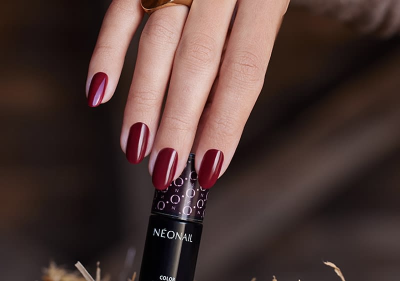 Hand mit dunkelrotem Nagellack auf einer schwarzen Nagellack-Flasche der Marke NEONAIL, Fingernägel oval geformt, Ring am Mittelfinger