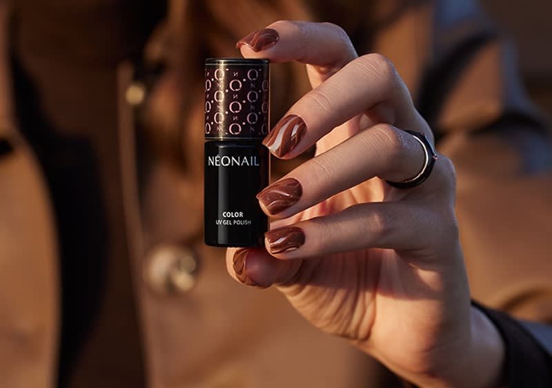 Hand hält Fläschchen Nagellack der Marke NEONAIL mit braunem, marmoriertem Gelnagellack an den Fingern, Hand trägt schwarzen Ring und braunen Mantel im Hintergrund