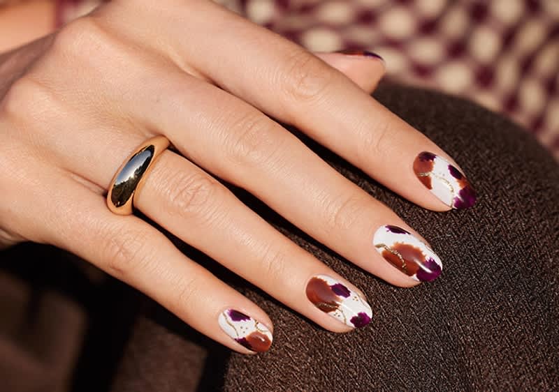 Hand mit goldfarbenem Ring auf Mittelfinger und weiß lackierten Fingernägeln mit braunen und violetten Farbflecken, Hand liegt auf dunkelbrauner Stoffoberfläche