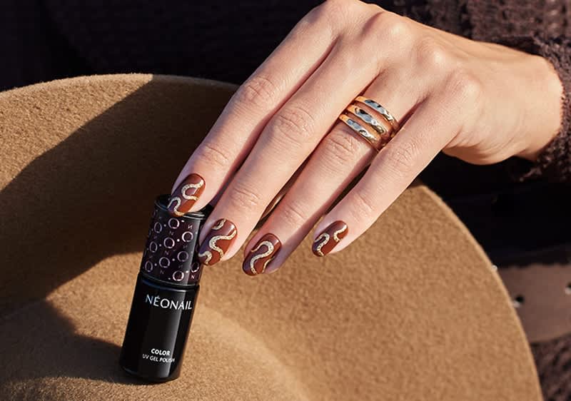 Hand mit goldfarbenen Ringen und braunem Nageldesign mit goldenen Linien hält schwarze Neónail Gel-Polish-Flasche auf hellbraunem Hut