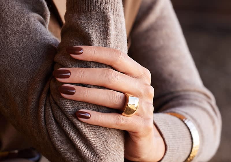 Zwei verschränkte Arme in beigem Strickpullover, eine Hand mit braun lackierten Nägeln trägt einen breiten goldenen Ring und ein goldfarbenes Armband