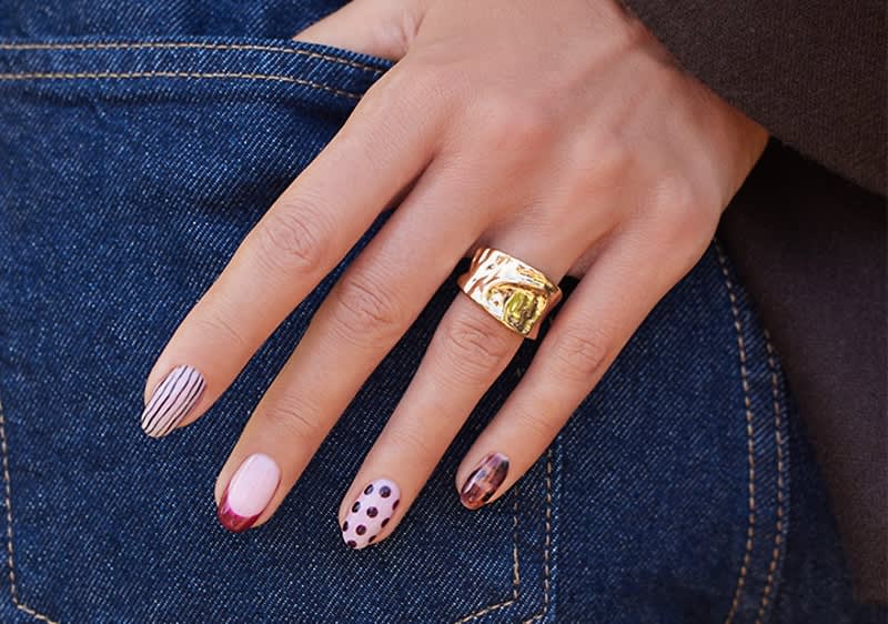 Hand mit kunstvoll lackierten Fingernägeln auf Jeanshose, Nägel mit unterschiedlichen Designs: rosé mit schwarzen Punkten, rosé mit dünnen schwarzen Streifen, dunkelroter French-Spitze und marmoriertes Muster, breiter goldener Ring am Mittelfinger