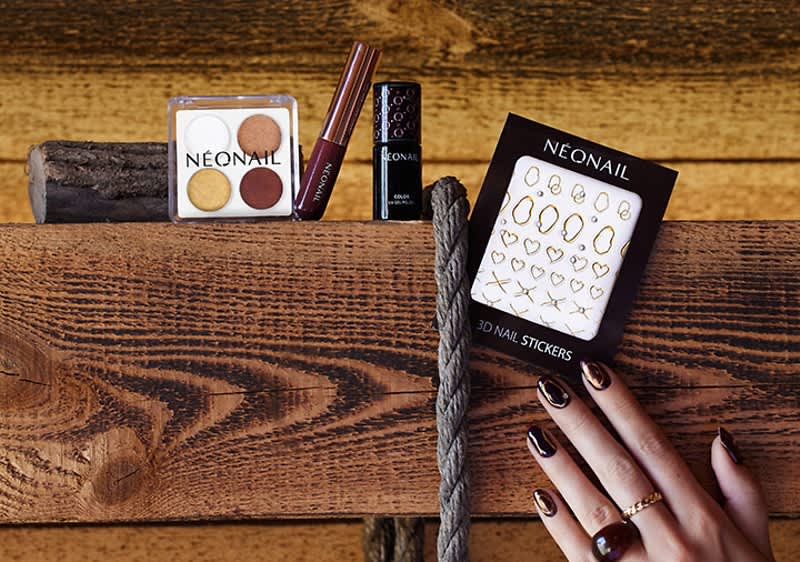 Vier Kosmetikprodukte der Marke Néonail auf Holz: Lidschattenpalette mit vier Tönen, dunkler Nagellack, braun-roter Lippenstiftstift und Packung mit goldenen 3D-Nagelstickern, rechts Hand mit dunkel lackierten, gemusterten Nägeln und Ringen auf dem Holzablagebrett