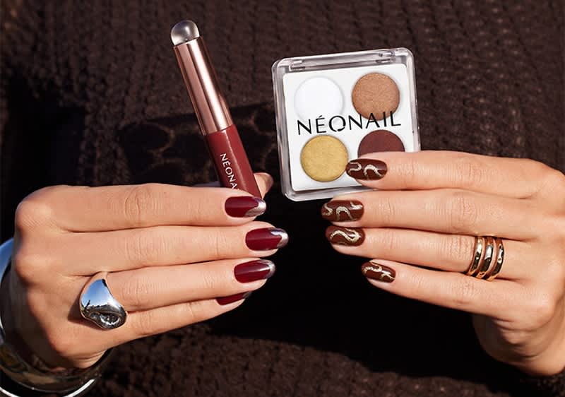 Hände mit dunkelrotem und braunem Nagellack, linke Hand hält roten Nagellackstift, rechte Hand hält eckige Lidschattenpalette mit Aufschrift „NEONAIL“ und vier Farbfeldern, Fingernägel teils mit goldenen Musterakzenten, Schmuckringe sichtbar