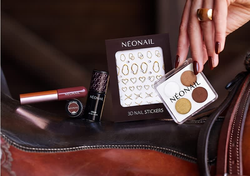 Mehrere Nagelprodukte der Marke NEONAIL auf Lederuntergrund: links Nagellack und Lippenstift, in der Mitte Packung mit 3D-Nail-Stickern, rechts offenes kleines Quad mit vier Lidschatten- oder Nagelfarben, Hand mit lackierten Fingern berührt das Quad