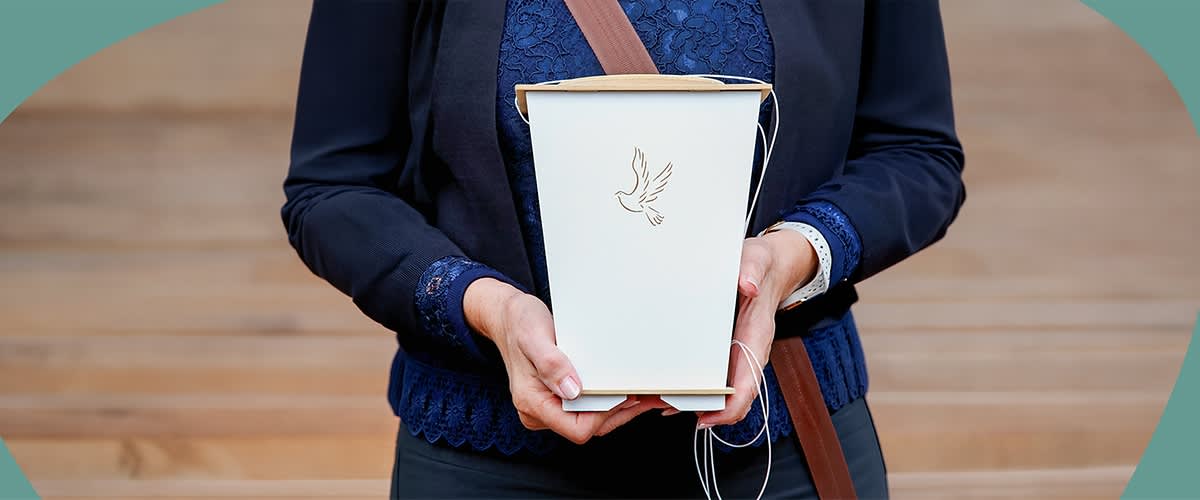 Person hält vor dem Oberkörper eine weiße Urnenbox mit goldfarbener Taubenzeichnung, Kleidung: dunkle Jacke und blaues Spitzenoberteil