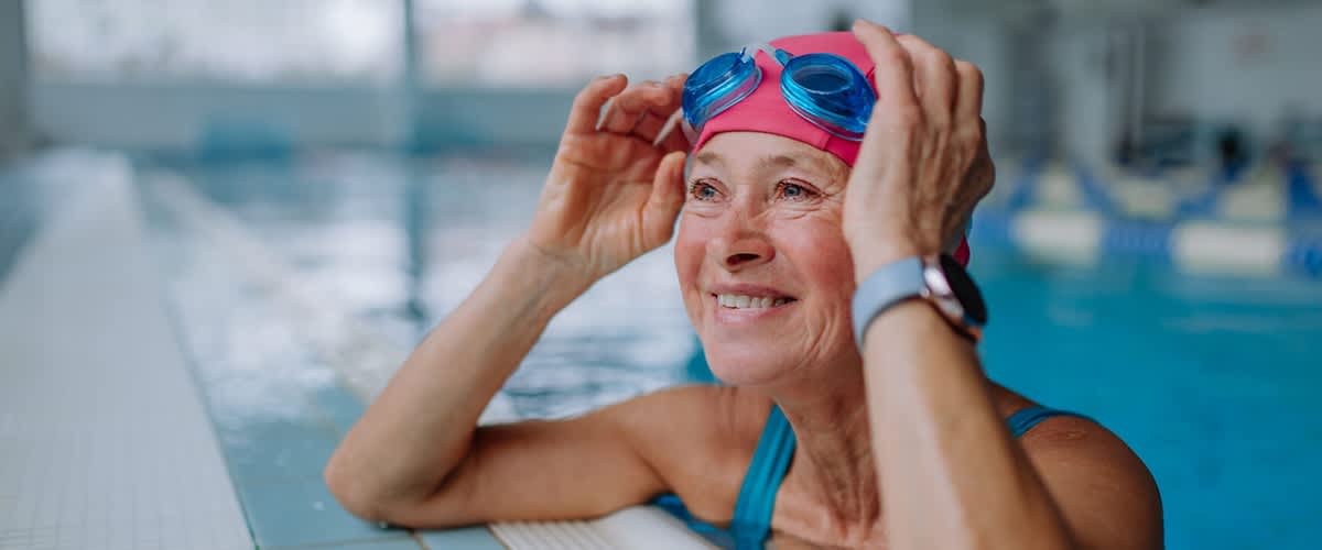 Ältere Frau am Schwimmbeckenrand mit rosa Badekappe und blauen Schwimmbrillen, sie lächelt und richtet die Brille mit den Händen, über dem Beckenrand sichtbar ein Fitnessarmband