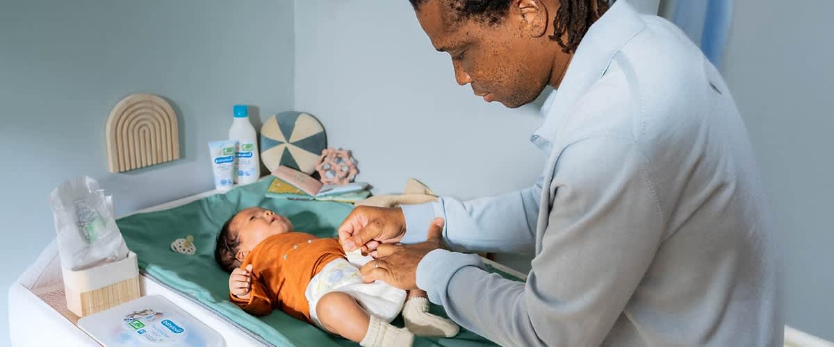 Ein Baby liegt auf einer Wickelunterlage und trägt einen orangefarbenen Pullover sowie weiße Socken, während eine Person mit dunkler Hautfarbe und lockigem Haar, deren Gesicht unscharf ist, die Windel des Babys wechselt; im Hintergrund sind Baby-Pflegeprodukte, ein Spielzeug und eine dekorative Wandgestaltung zu sehen.