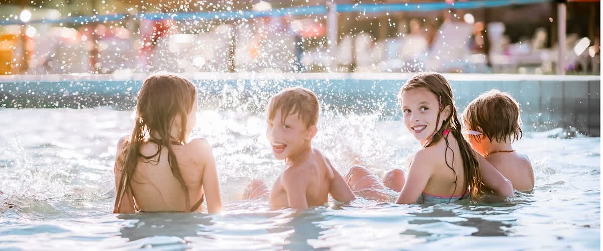 Vier Kinder von hinten und seitlich im Schwimmbecken, zwei in der Bildmitte spritzend und lachend, im Hintergrund Sonnenschirme und Badende am Beckenrand