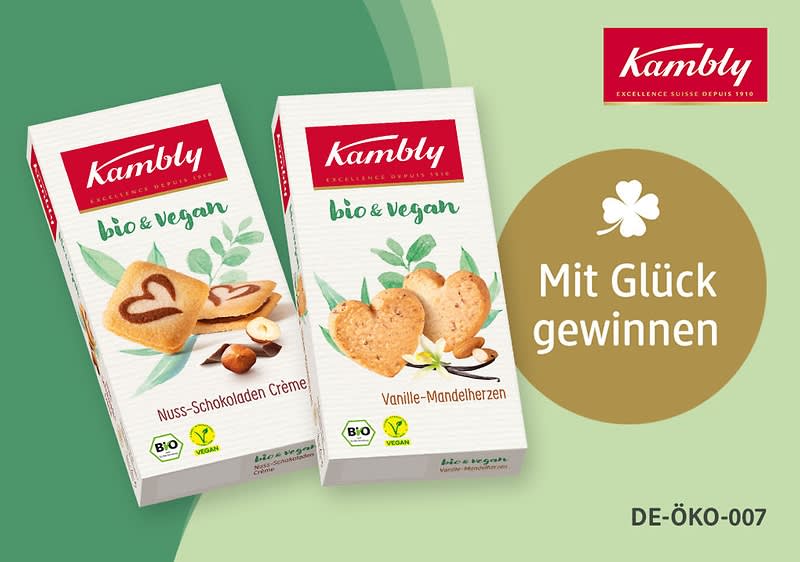 Zwei Kambly Bio & Vegan Keks-Schachteln nebeneinander: links „Nuss-Schokoladen Crème“ mit Herzkeksen und Haselnuss, rechts „Vanille-Mandelherzen“ mit herzförmigen Keksen und Mandeln; rechts ein goldener Kreis mit Text „Mit Glück gewinnen“ und oben rechts rotes Kambly-Logo