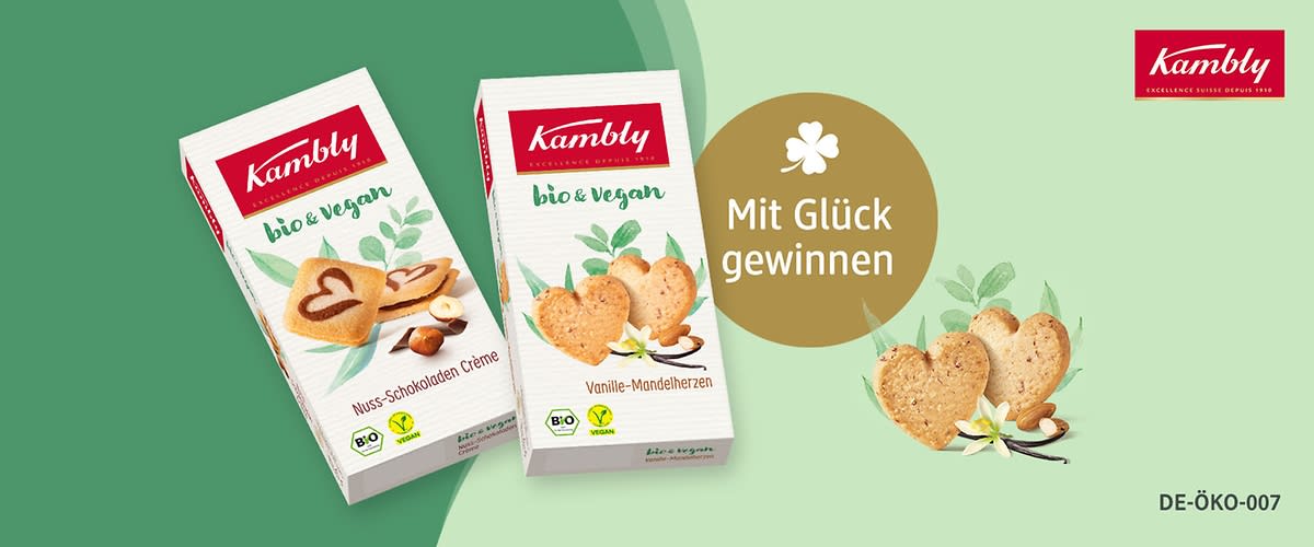 Zwei Kambly Bio & Vegan Keks-Schachteln nebeneinander: links „Nuss-Schokoladen Crème“ mit Herzkeksen und Haselnuss, rechts „Vanille-Mandelherzen“ mit herzförmigen Keksen und Mandeln; rechts ein goldener Kreis mit Text „Mit Glück gewinnen“ und oben rechts rotes Kambly-Logo