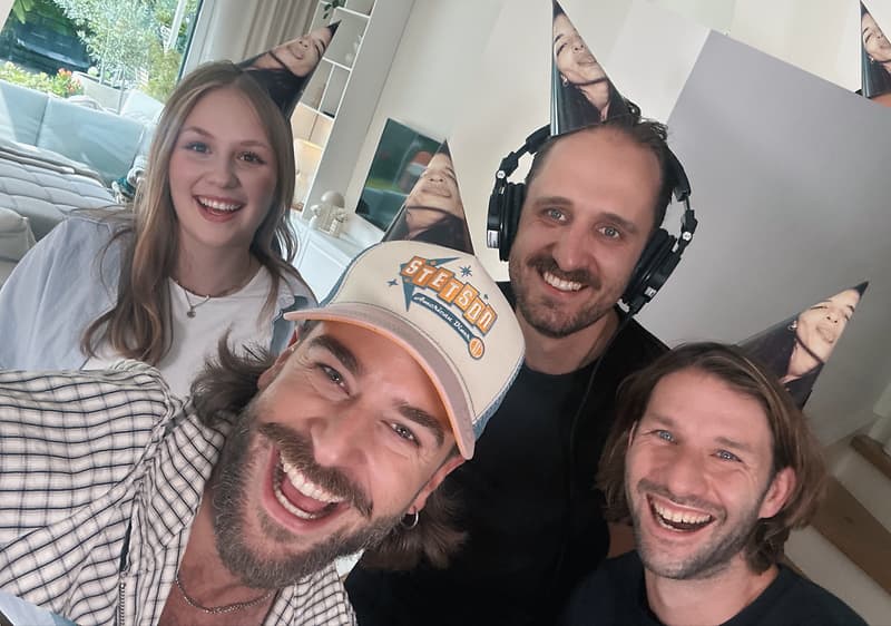 Vier Erwachsene mit Partyhüten stehen dicht beieinander und lächeln in die Kamera, eine Person trägt Kopfhörer und eine weitere eine hellblaue Baseballkappe mit orange-blauem Schriftzug, im Hintergrund ein Wohnraum mit Sofa und Fenster