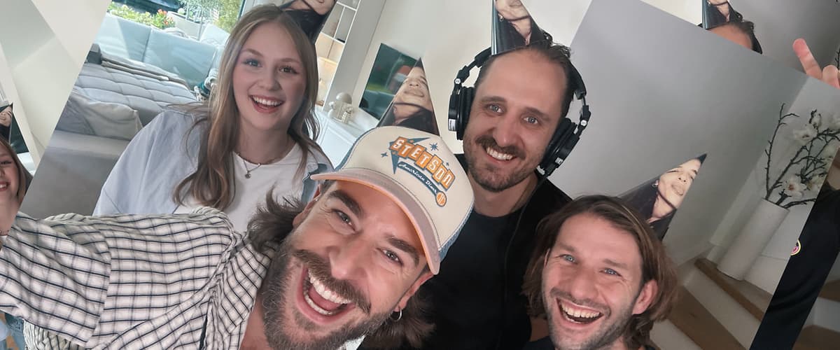 Vier Erwachsene mit Partyhüten stehen dicht beieinander und lächeln in die Kamera, eine Person trägt Kopfhörer und eine weitere eine hellblaue Baseballkappe mit orange-blauem Schriftzug, im Hintergrund ein Wohnraum mit Sofa und Fenster