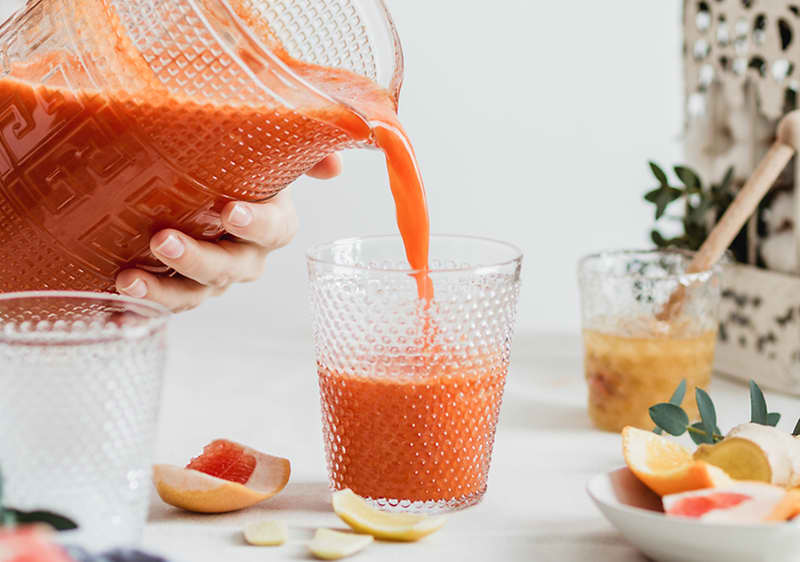 Jemand gießt roten Fruchtsaft aus einem Glas-Krug in ein strukturiertes Glas, im Vordergrund liegen Grapefruit- und Zitronenscheiben und im Hintergrund ein Glas mit Honig oder Marmelade und ein kleines Pflanzenblatt