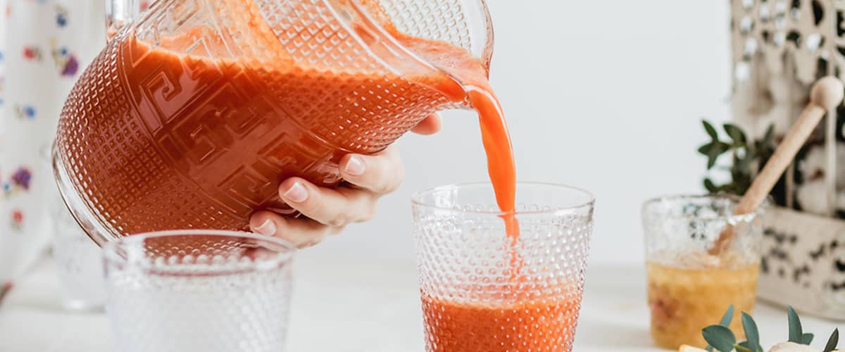 Jemand gießt roten Fruchtsaft aus einem Glas-Krug in ein strukturiertes Glas, im Vordergrund liegen Grapefruit- und Zitronenscheiben und im Hintergrund ein Glas mit Honig oder Marmelade und ein kleines Pflanzenblatt