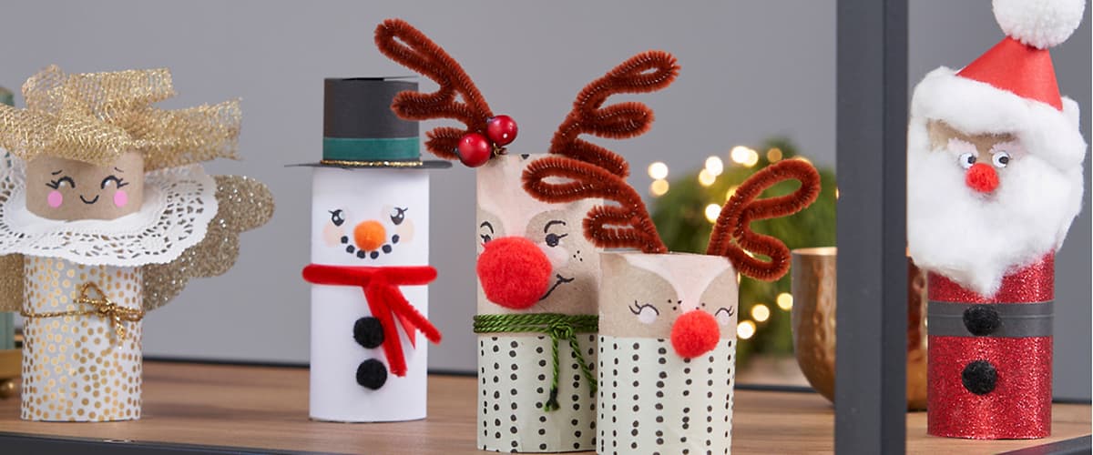 Drei Weihnachtspuppen aus bemalten Toilettenpapierrollen: links ein Schneemann mit schwarzem Zylinder, rotem Schal, Karottennase und Knopfreihen, rechts zwei Rentiere mit roten Pompomnasen, braunen Pfeifenputzer-Geweihen und grünen Bändern, im Hintergrund Lichterdekoration
