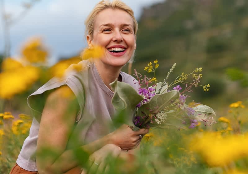 Frau kniet in einem Blumenfeld und hält einen spontanen Strauß aus Wildblumen, sie lächelt und schaut nach rechts