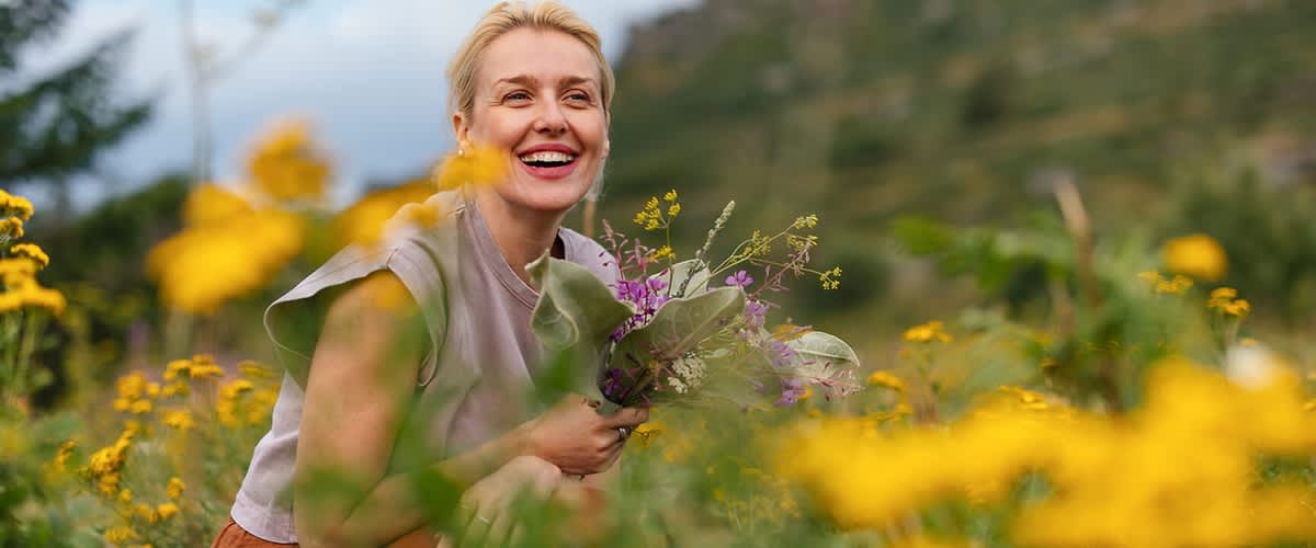 Frau kniet in einem Blumenfeld und hält einen spontanen Strauß aus Wildblumen, sie lächelt und schaut nach rechts