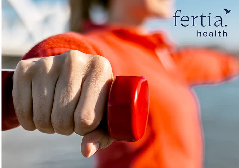 Hand hält ein kleines rotes Hantelgewicht ausgestreckt, im Hintergrund unscharf Person in orangenem Oberteil, oben rechts Markenname „fertia health“