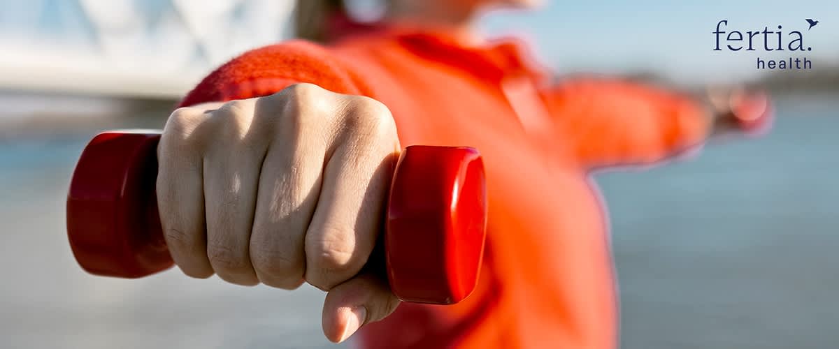 Hand hält ein kleines rotes Hantelgewicht ausgestreckt, im Hintergrund unscharf Person in orangenem Oberteil, oben rechts Markenname „fertia health“