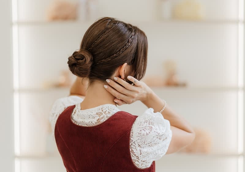 Low Bun: Frau von hinten mit hochgestecktem Dutt und geflochtener Strähne, trägt weiße Spitzenbluse unter ärmellosem, dunkelrotem Kleid und hält Hände an den Nacken ohne Blick zur Kamera