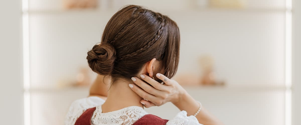Low Bun: Frau von hinten mit hochgestecktem Dutt und geflochtener Strähne, trägt weiße Spitzenbluse unter ärmellosem, dunkelrotem Kleid und hält Hände an den Nacken ohne Blick zur Kamera