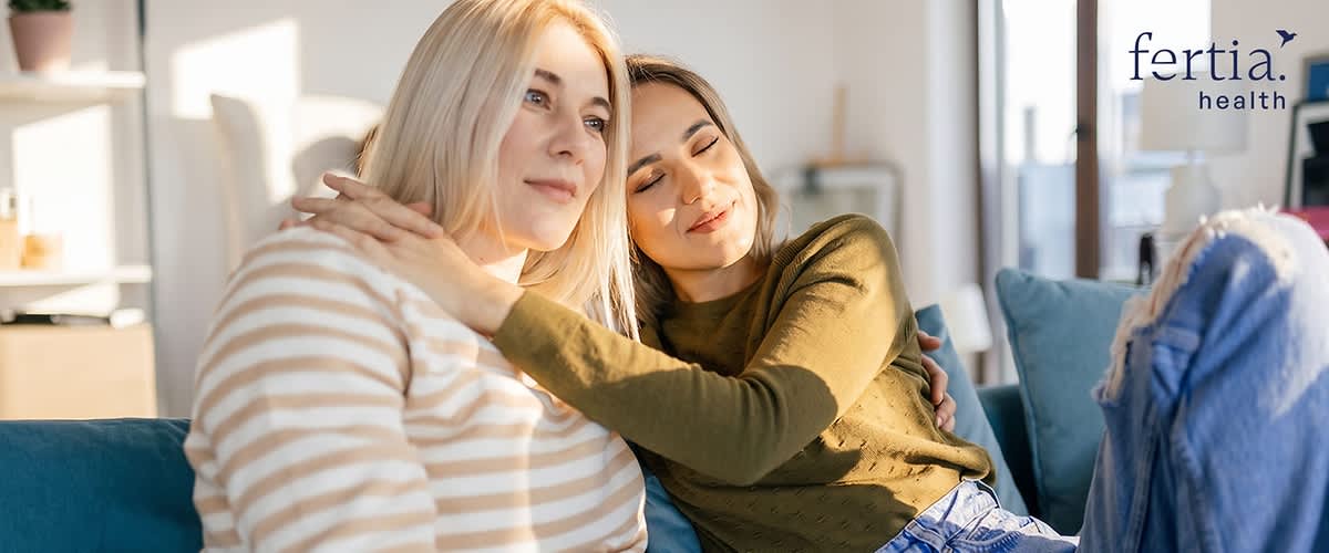 Zwei auf einem Sofa sitzende Frauen, die eine umarmt die andere von der Seite