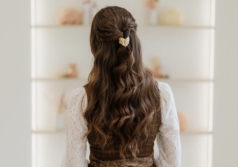 Frau von hinten mit langen gewellten braunen Haaren, halb hochgesteckt und mit einer goldenen Herz-Haarklammer befestigt, sie trägt eine weiße Spitzenbluse und eine braune Weste
