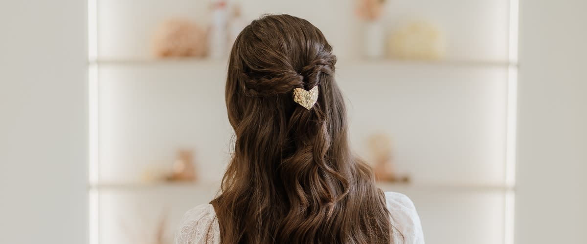 Frau von hinten mit langen gewellten braunen Haaren, halb hochgesteckt und mit einer goldenen Herz-Haarklammer befestigt, sie trägt eine weiße Spitzenbluse und eine braune Weste