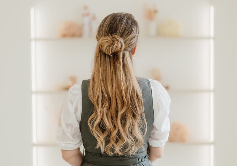 Rückansicht einer Frau mit langem, welligem blonden Haar, das zu einem lockeren Knoten halb hochgesteckt ist, sie trägt ein weißes Oberteil und ein dunkelgrünes Kleid oder eine Schürze