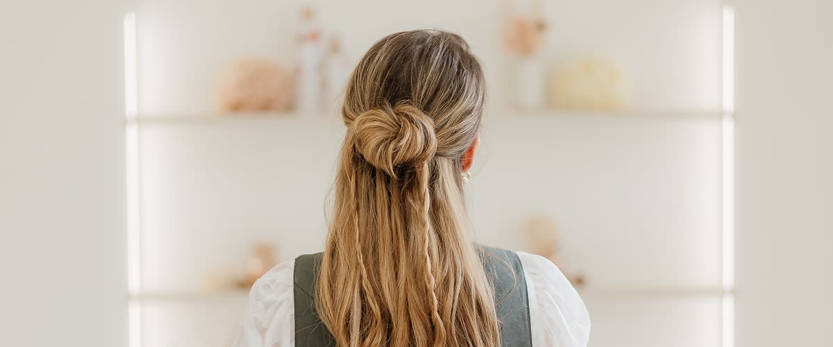 Rückansicht einer Frau mit langem, welligem blonden Haar, das zu einem lockeren Knoten halb hochgesteckt ist, sie trägt ein weißes Oberteil und ein dunkelgrünes Kleid oder eine Schürze
