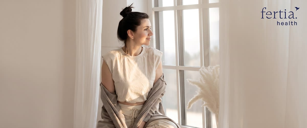Frau sitzt entspannt auf einer Fensterbank, trägt helle Loungewear und einen offenen Cardigan, Blick nach rechts aus dem Bild