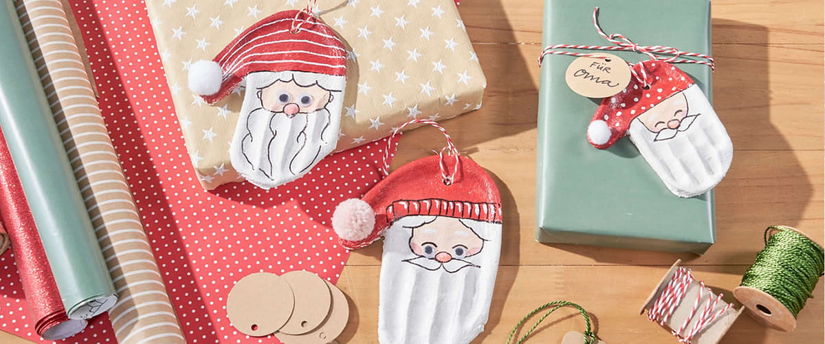 Drei handgemachte Weihnachtsanhänger in Form von Weihnachtsmannköpfen mit roter Mütze und weißem Bart auf Geschenkpapier und Holztisch, dazu runde und herzförmige Geschenkanhänger aus Kraftpapier