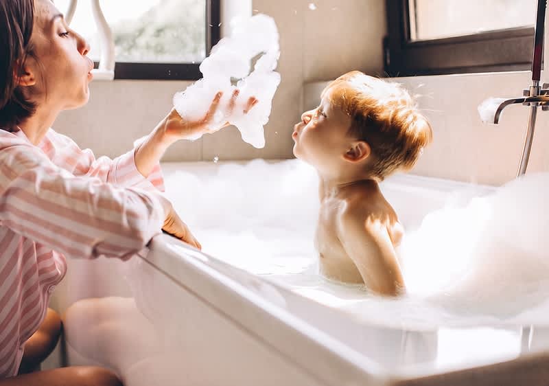 Mutter kniet neben einer Badewanne und formt Schaum, während ein Kleinkind im Schaum sitzt und den Schaum anbläst