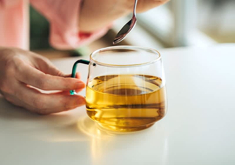 Hände halten eine Glas-Tasse mit hellem Tee, über der ein Löffel gehalten wird, der Tee berührt werden könnte