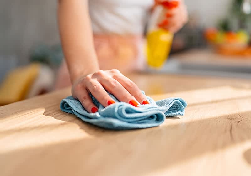 Hand mit roten Fingernägeln wischt eine Holzarbeitsfläche mit blauem Mikrofasertuch, im Hintergrund unscharf Sprühflasche sichtbar