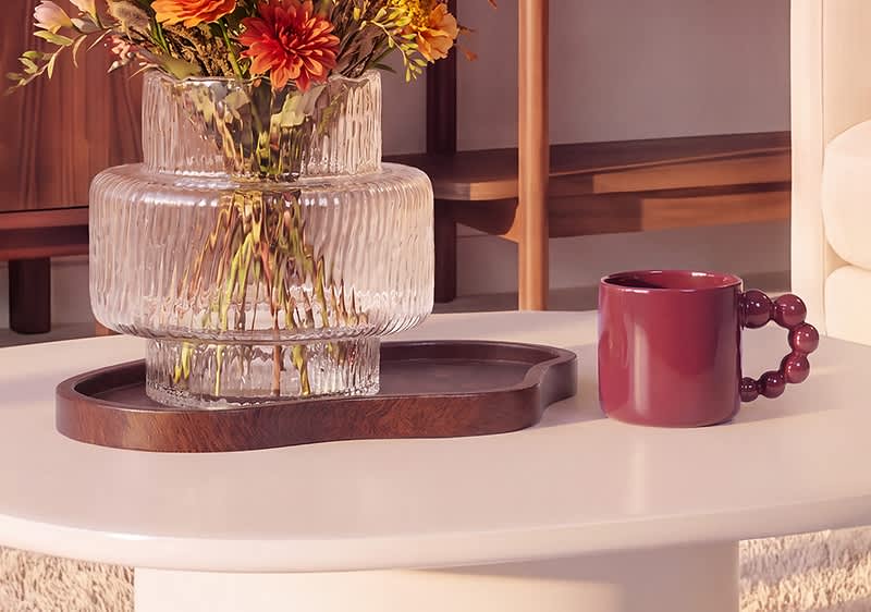 Transparente, geriffelte Glasvase mit bunten Herbstblumen auf einem dunklen Holztablett, daneben eine rotbraune Keramiktasse mit auffälligem Perlenhenkel. Bild mithilfe von KI erstellt