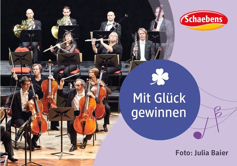 Orchester mit Streichern und Bläsern links, rechts lila Flächen mit blauem Kreis und weißer Schrift „Mit Glück gewinnen“, oben rechts rotes Schaebens-Logo und unten rechts Fotocredit „Foto: Julia Baier“