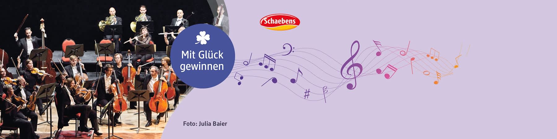 Orchester mit Streichern und Bläsern auf der Bühne links, rechts lila Bereich mit rundem blauen Kreis und weißer Schrift „Mit Glück gewinnen“, oben rechts rotes Schaebens-Logo, unten rechts Fotoangabe „Foto: Julia Baier“