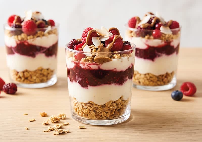 Drei Gläser mit Schichtdessert aus Granola, Joghurt, roter Beerenschicht, Nüssen und frischen Beeren, mittleres Glas im Vordergrund ohne abschließenden Punkt