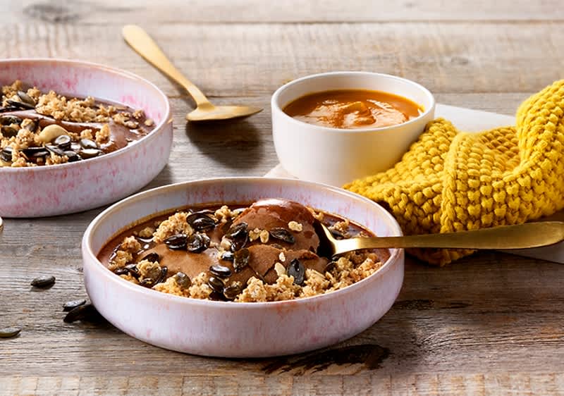 Zwei flache Schalen mit Schokoladenpudding, Hafer-Crunch und Kürbiskernen auf Holztisch, daneben eine kleine weiße Schale mit orangefarbener Sauce und ein gelbes gestricktes Tuch