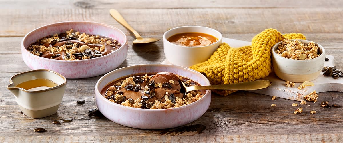 Zwei flache Schalen mit Schokoladenpudding, Hafer-Crunch und Kürbiskernen auf Holztisch, daneben eine kleine weiße Schale mit orangefarbener Sauce und ein gelbes gestricktes Tuch