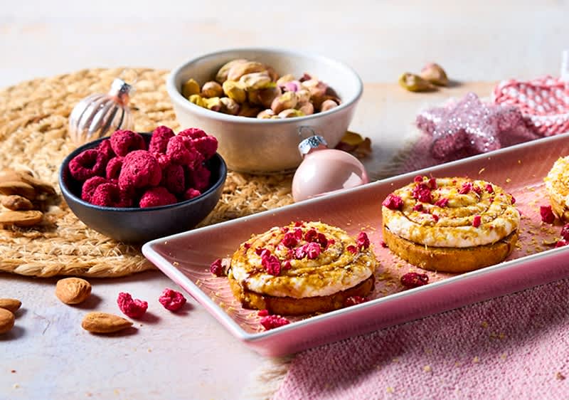 Drei kleine Creme-Törtchen mit rosa Fruchtstückchen auf einem länglichen rosa Teller, daneben Schalen mit gefriergetrockneten Himbeeren und Pistazien sowie verstreute Mandeln und Weihnachtskugeln