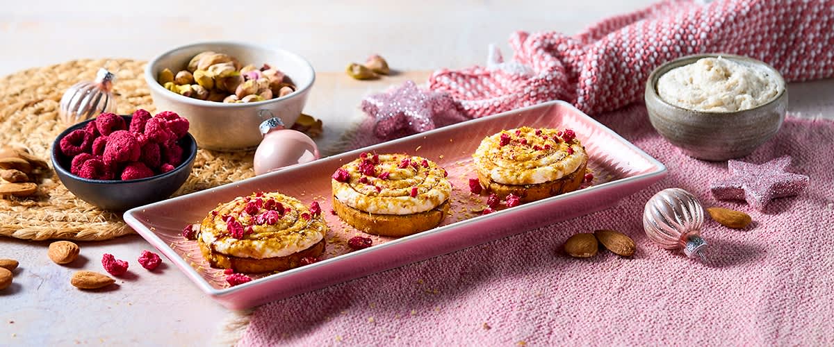 Drei kleine Creme-Törtchen mit rosa Fruchtstückchen auf einem länglichen rosa Teller, daneben Schalen mit gefriergetrockneten Himbeeren und Pistazien sowie verstreute Mandeln und Weihnachtskugeln