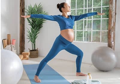 Eine schwangere Frau in blauer Sportkleidung steht auf einer blauen Yogamatte und macht eine Yoga-Übung in einem hellen Raum mit großen Fenstern, im Hintergrund sind ein großer Gymnastikball, eine Pflanze in einem Korb und mehrere zusammengerollte Yogamatten zu sehen, daneben steht eine Flasche mit Lotion auf dem Boden