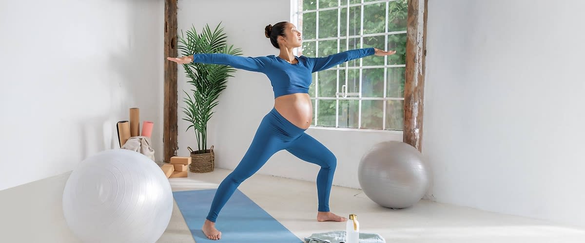 Eine schwangere Frau in blauer Sportkleidung steht auf einer blauen Yogamatte und macht eine Yoga-Übung in einem hellen Raum mit großen Fenstern, im Hintergrund sind ein großer Gymnastikball, eine Pflanze in einem Korb und mehrere zusammengerollte Yogamatten zu sehen, daneben steht eine Flasche mit Lotion auf dem Boden