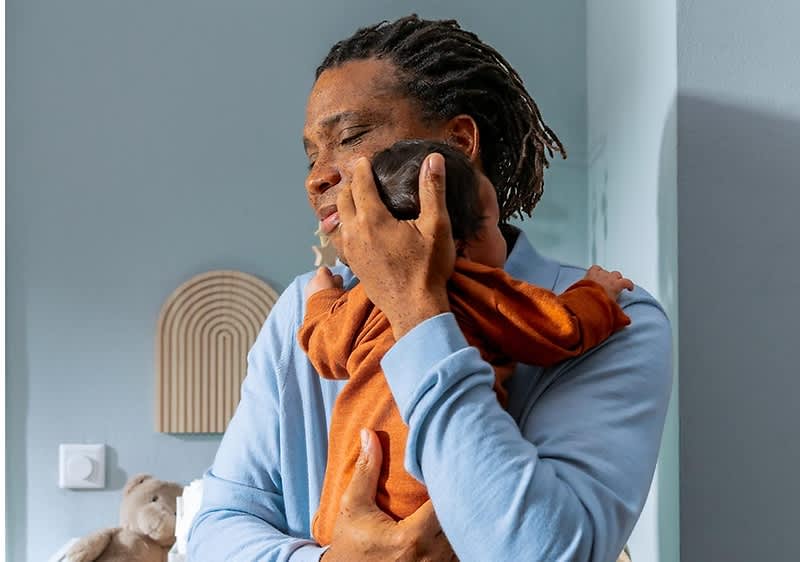 Ein Mann mit Dreadlocks trägt ein Baby in einem orangefarbenen Strampler auf dem Arm, während er in einem Raum mit hellblauen Wänden steht, im Hintergrund sind ein dekoratives Wandobjekt in Form eines Halbkreises und ein Plüschhase auf einem Möbelstück zu sehen