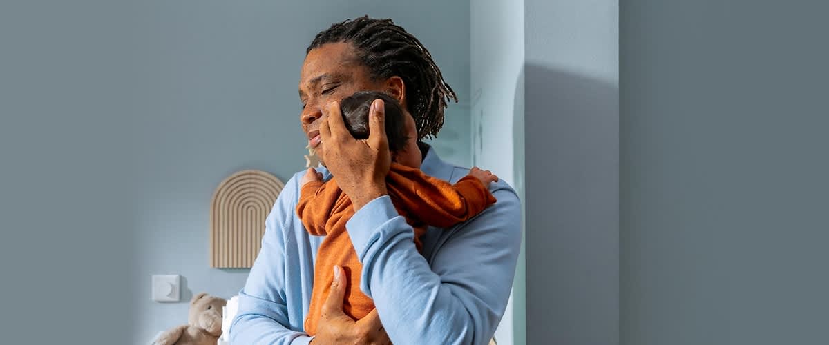 Ein Mann mit Dreadlocks trägt ein Baby in einem orangefarbenen Strampler auf dem Arm, während er in einem Raum mit hellblauen Wänden steht, im Hintergrund sind ein dekoratives Wandobjekt in Form eines Halbkreises und ein Plüschhase auf einem Möbelstück zu sehen